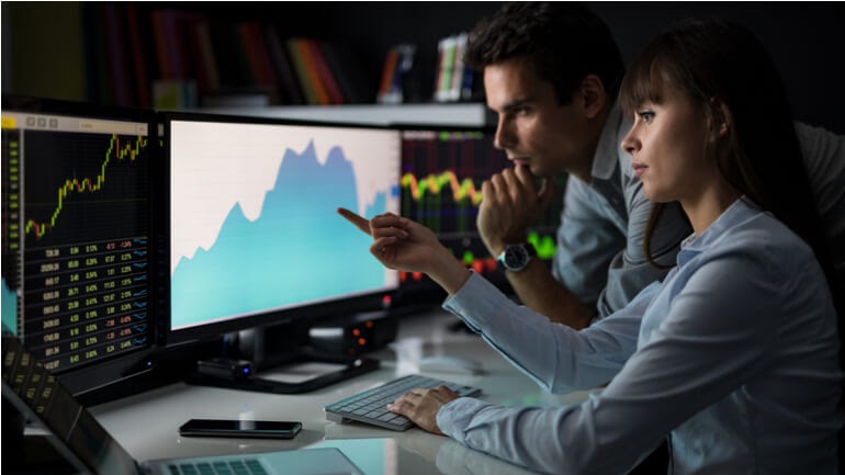 Sfondo di un ufficio con un pc e due persone che indicano sul pc un grafico a linee e uno candlestick trading a indicare il market sentiment nel trading.