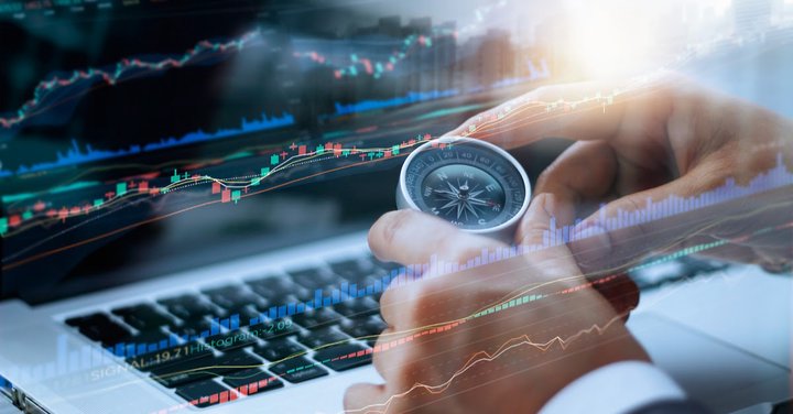 Sfondo di un computer portatile con grafico candlestick e altri tipi di grafici trading sullo schermo, e una mano che tiene una bussola, a indicare l'analisi fondamentale.