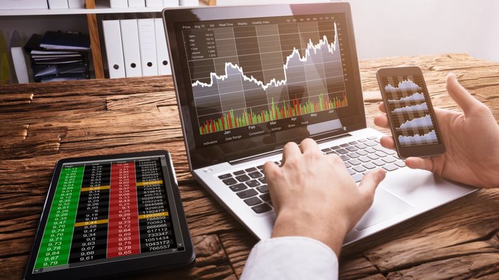 Sfondo di ufficio con persona che ha un portatile e un cellulare alla mano ed entrambi mostrano nei rispettivi schermi un grafico di trading, per indicare come inziiare a fare trading.