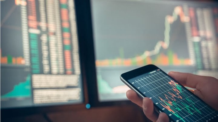 Sfondo di schermi con grafici trading candlestick e a linee e mano con schermo di cellulare con grafico trading a linee,  a indicare l'investimento in strumenti derivati finanziari.