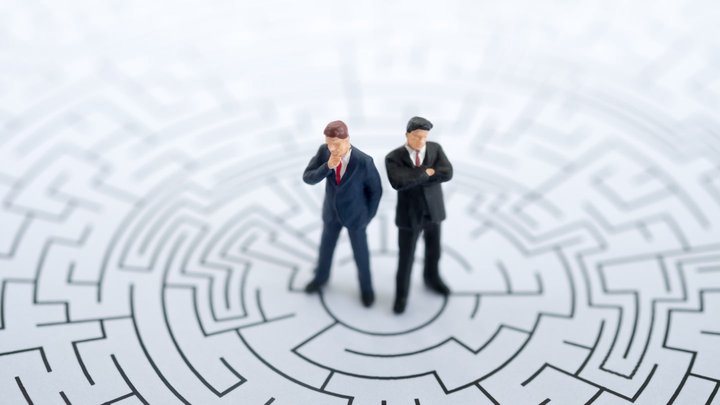 Sfondo bianco con pavimento a labirinto e due uomini identici con espressione pensante o rassegnata per indicare la psicologia del trading.