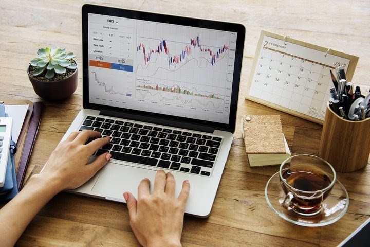 Sfondo di un tavolo con computer portatile che mostra la schermata di una piattaforma trading con grafico candlestick e schermata nuovo ordine, a indicare le diverse strategie investimento.